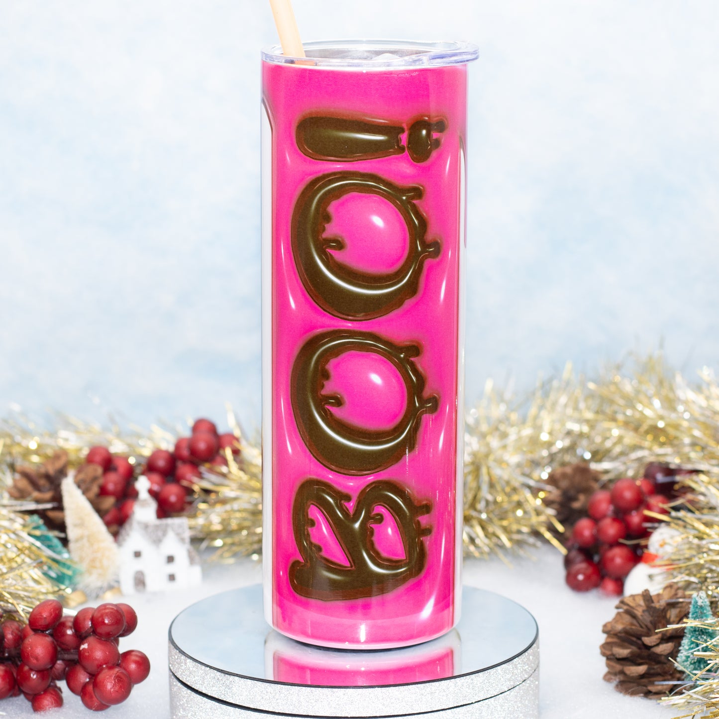 A tall Stainless Steel tumbler with a bright pink insert and large brown 'BOO' lettering, shown on a silver pedestal with holiday tinsel, berries, and a straw against a pale blue background.