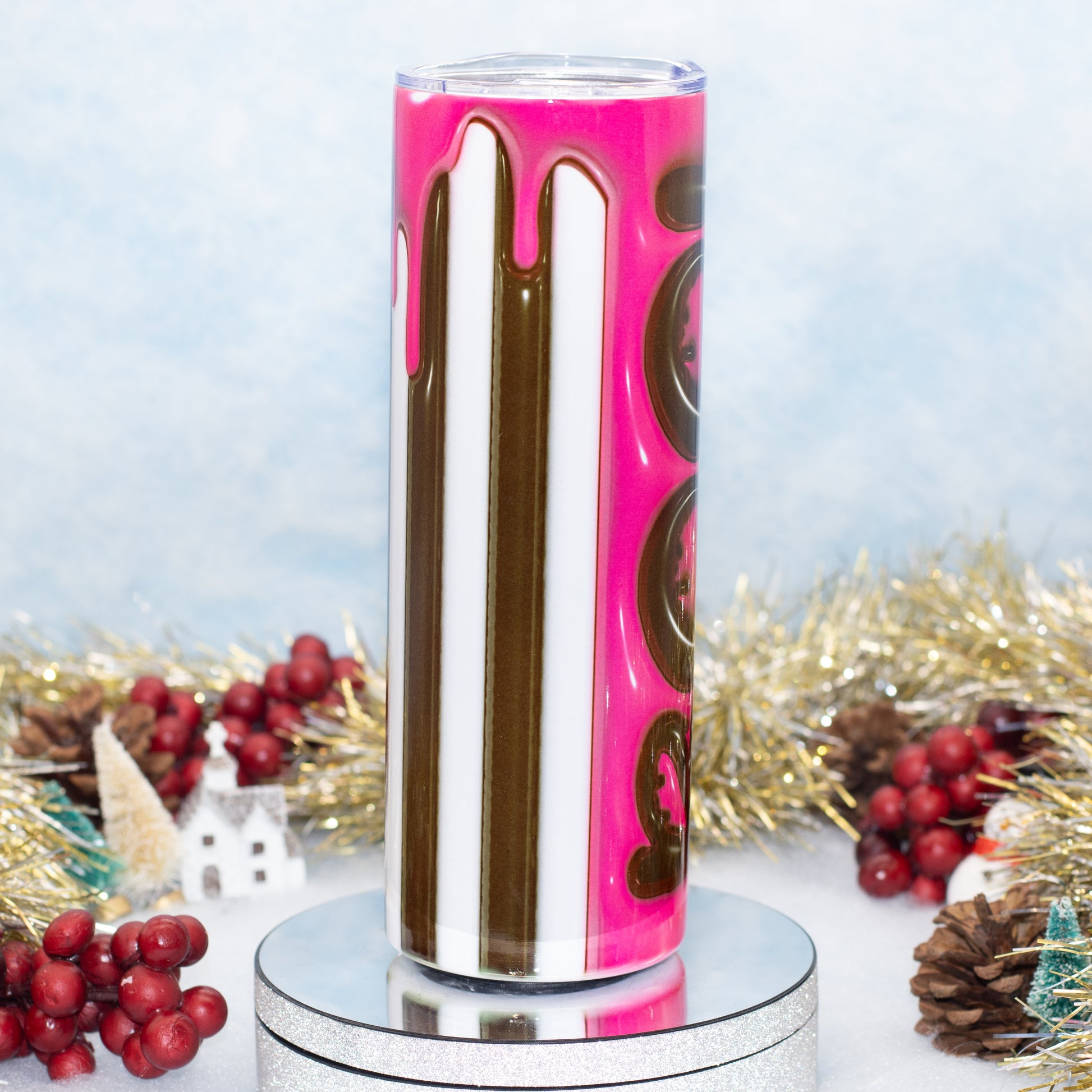 A cylindrical tumbler with a bright pink and white vertical stripe design, accented by dripping pink and brown graphics, displayed on a silver pedestal with holiday decorations in the background.