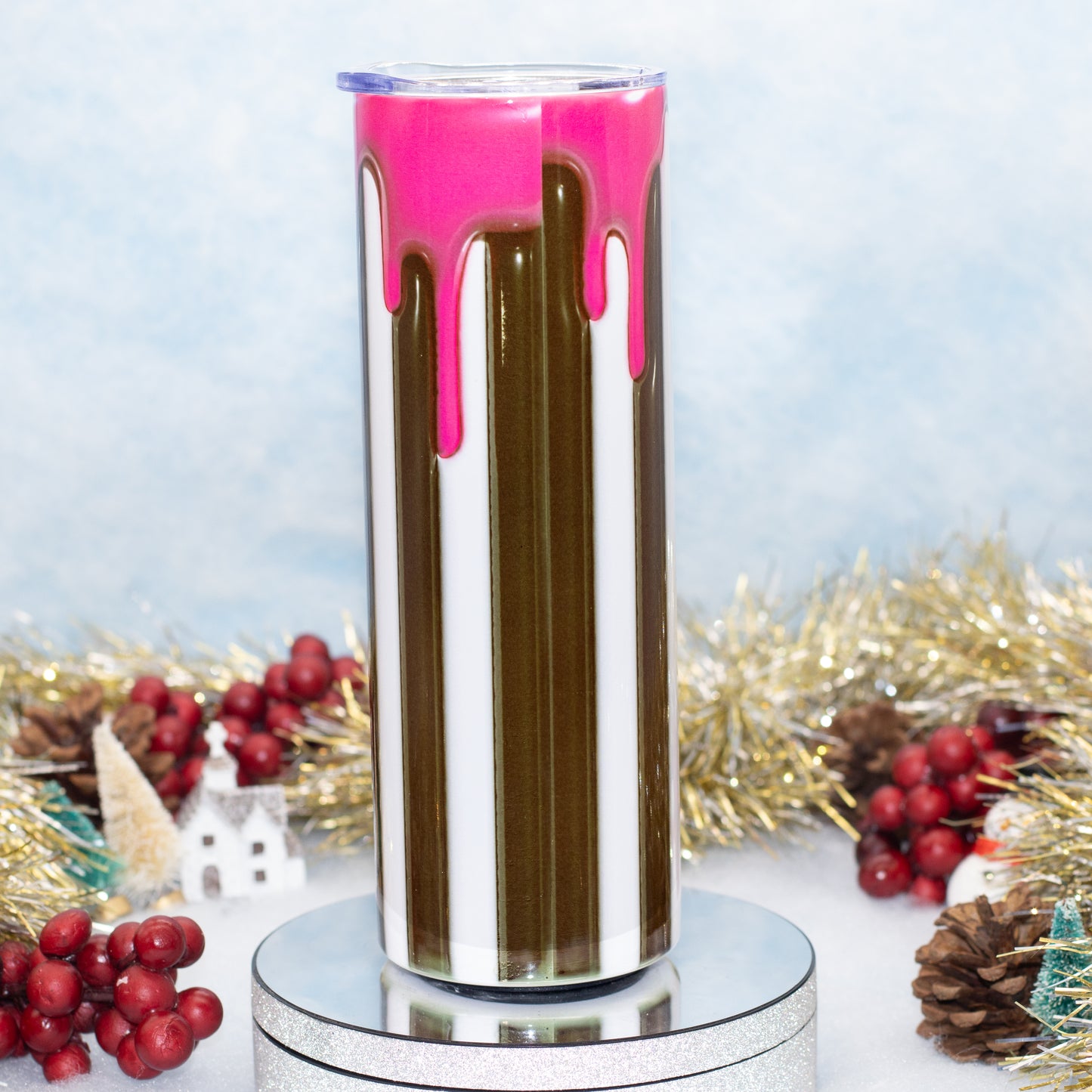 A tall insulated tumbler with a clear lid, featuring vertical stripes of white, dark brown, and bright pink dripping paint, displayed on a silver pedestal with holiday decorations in the background.