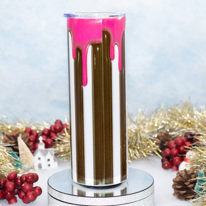 A tall insulated tumbler with a clear lid, featuring vertical stripes of white, dark brown, and bright pink dripping paint, displayed on a silver pedestal with holiday decorations in the background.