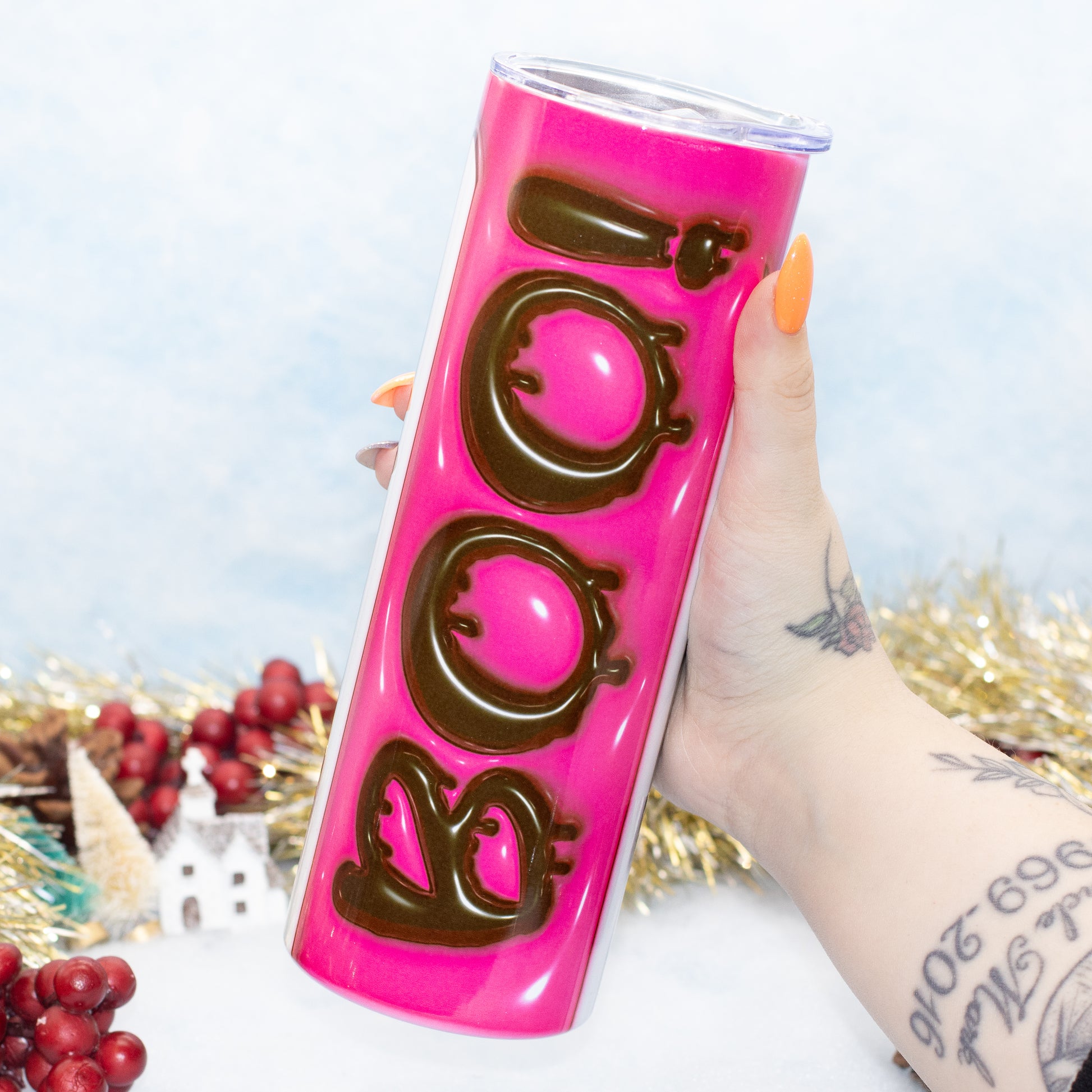 tumbler featuring a bright hot pink background and large dark brown 3D letters spelling 'BOO'. The background is decorated with gold tinsel, red berries, and a miniature white house.