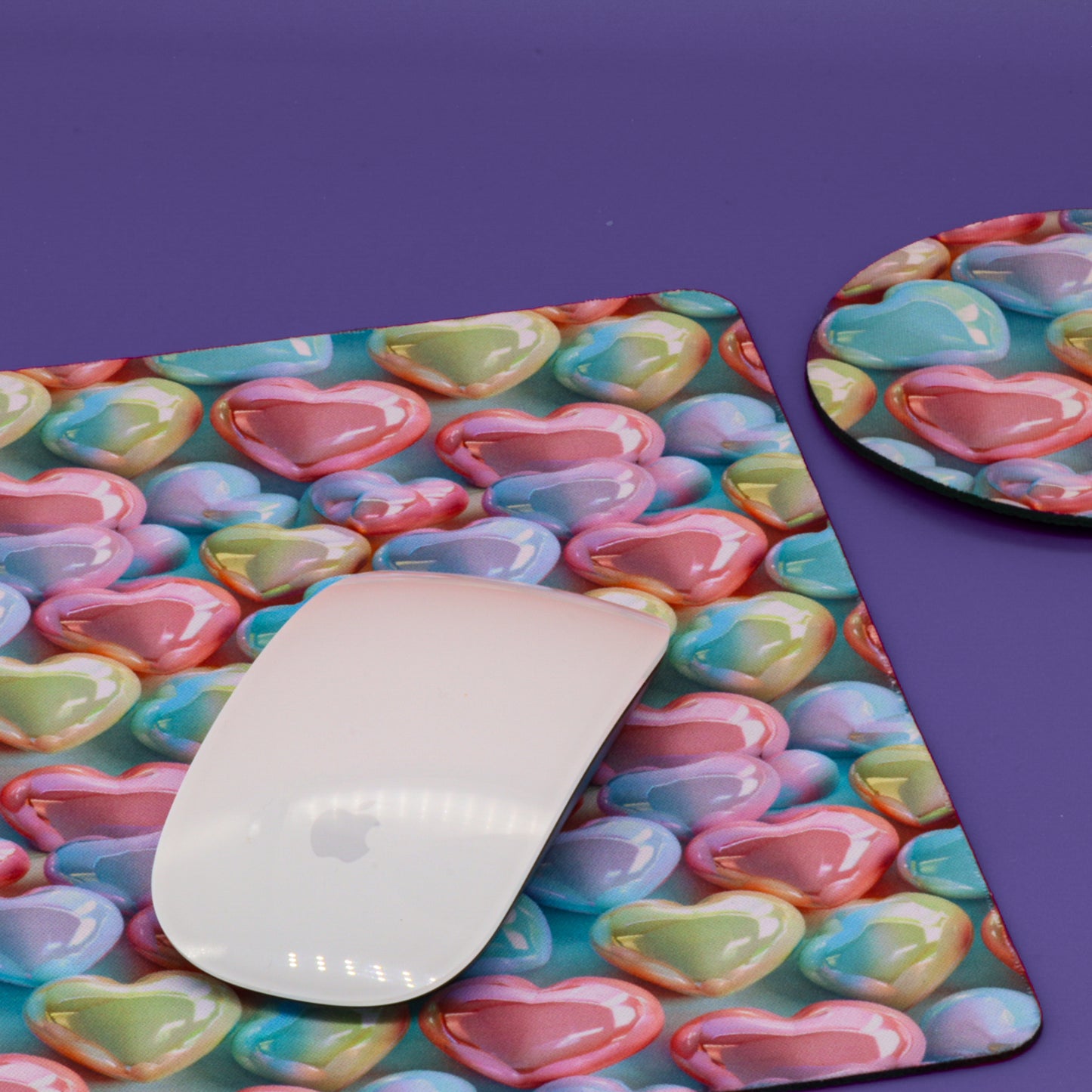 A rectangular desk mat and matching round coaster with a colorful heart pattern, shown on a purple background with a white computer mouse placed on the mat.