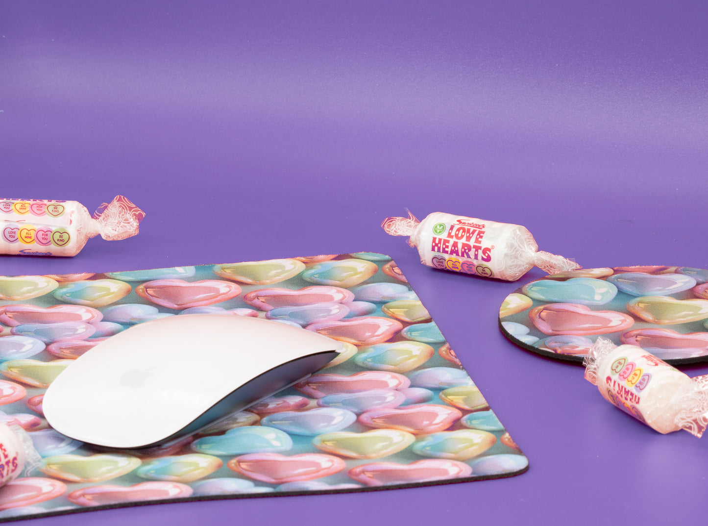 A desk setup featuring a large mouse pad and matching coasters with a colorful candy heart pattern, styled with a white wireless mouse and wrapped lollipops on a purple background.