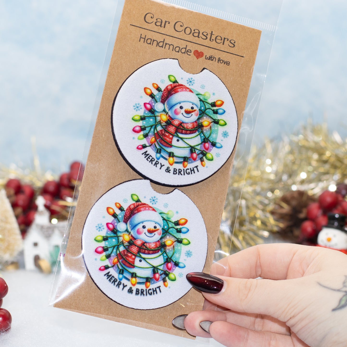 A hand holds a clear plastic package containing two round car coasters. The coasters feature a festive snowman illustration surrounded by colorful Christmas string lights on a white background, with the text 'MERRY & BRIGHT'.