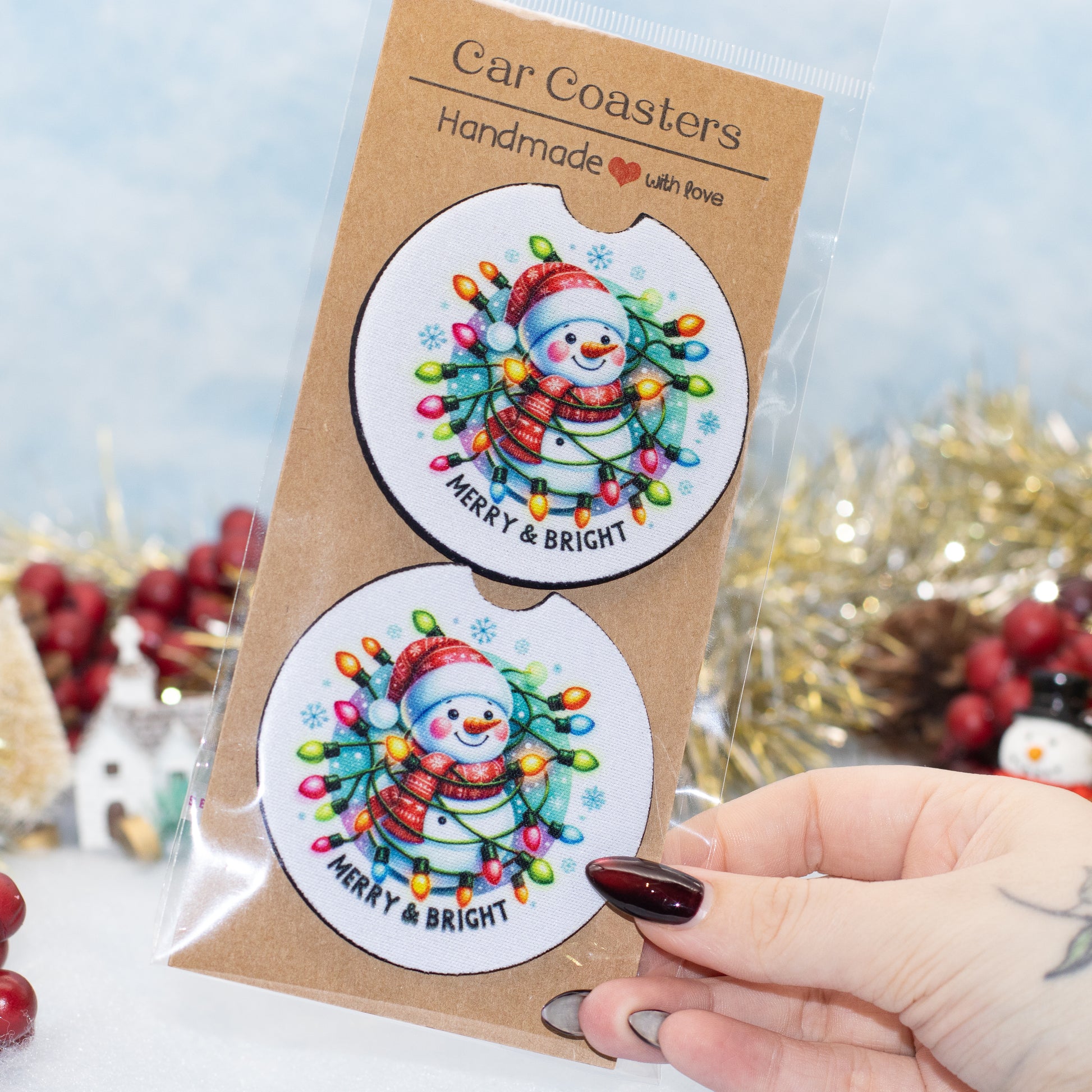 A hand holds a clear plastic package containing two round car coasters. The coasters feature a festive snowman illustration surrounded by colorful Christmas string lights on a white background, with the text 'MERRY & BRIGHT'.