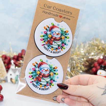 A hand holds a clear plastic package containing two round car coasters. The coasters feature a festive snowman illustration surrounded by colorful Christmas string lights on a white background, with the text 'MERRY & BRIGHT'.