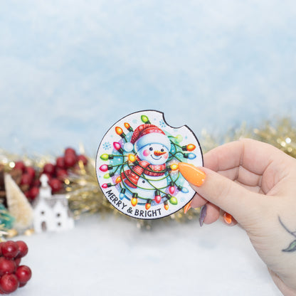 A hand holds a car coaster  featuring a snowman in a red hat and scarf surrounded by colourful holiday lights, with the text 'MERRY & BRIGHT' at the bottom. The background is a soft, out-of-focus light blue with gold tinsel and red berries.