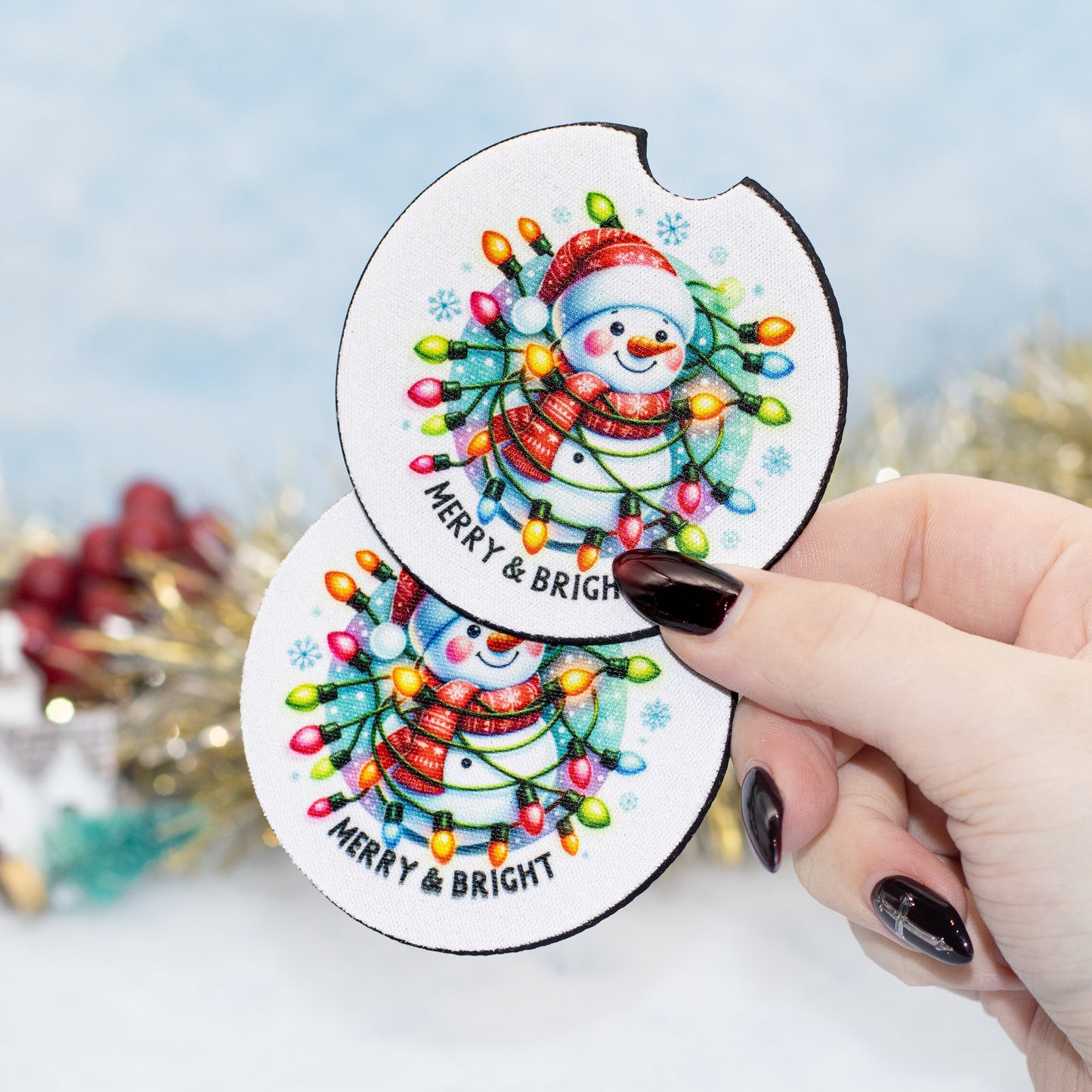 A hand holds two round white coasters featuring a colourful Christmas snowman design with glowing holiday lights and the text 'MERRY & BRIGHT'. The background is softly blurred with festive decorations.