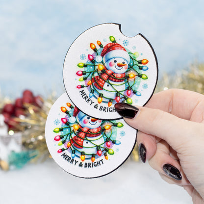 A hand holds two round white coasters featuring a colourful Christmas snowman design with glowing holiday lights and the text 'MERRY & BRIGHT'. The background is softly blurred with festive decorations.