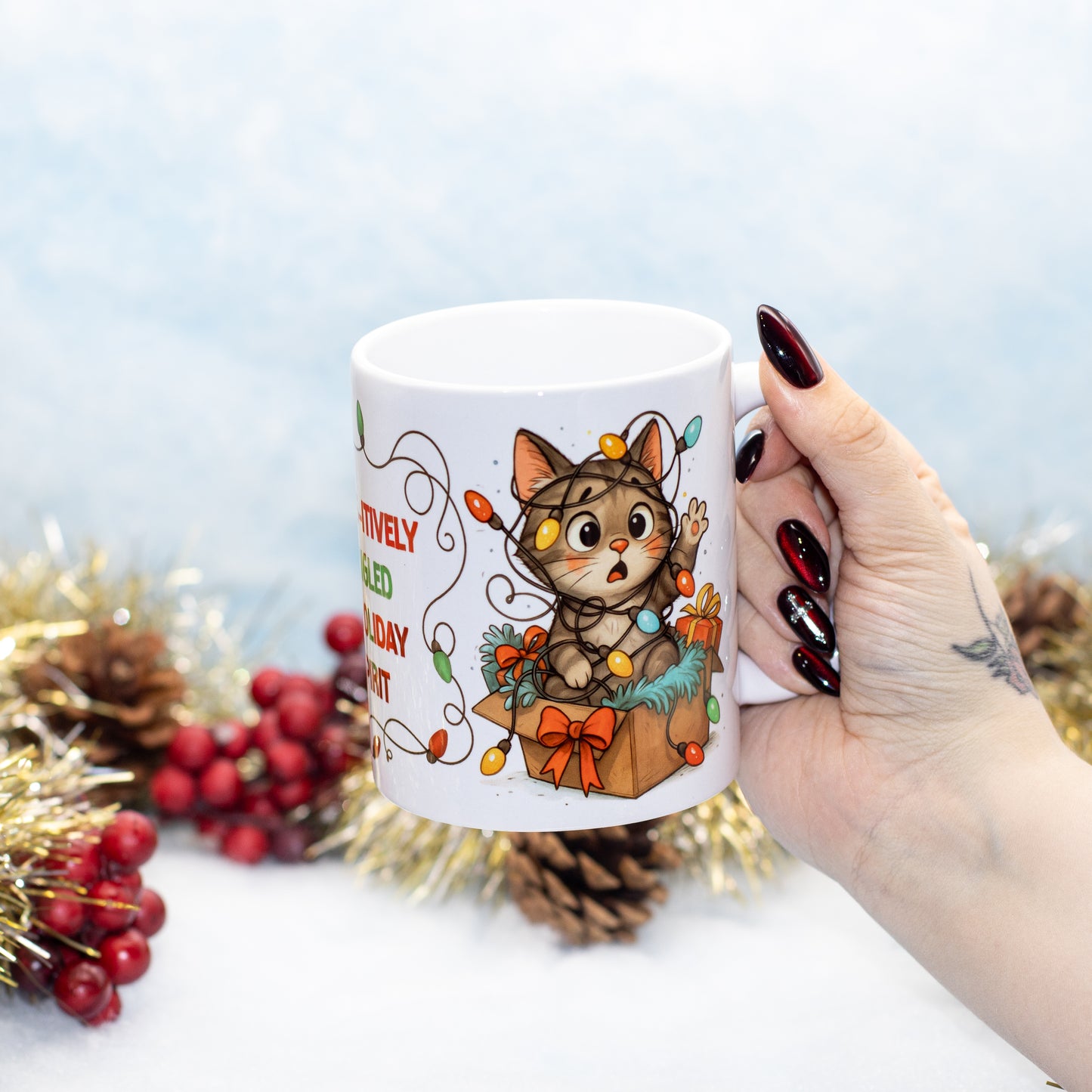 a white ceramic mug featuring a cute cartoon tabby cat sitting in a gift box surrounded by Christmas lights. The background is decorated with pinecones, red berries, and gold tinsel against a soft light blue backdrop.