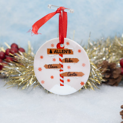 A round white Christmas ornament with a red ribbon hanger, featuring personalized names on wooden signpost-style labels and small red snowflake motifs, displayed on a snowy surface with tinsel and pinecones in the background.