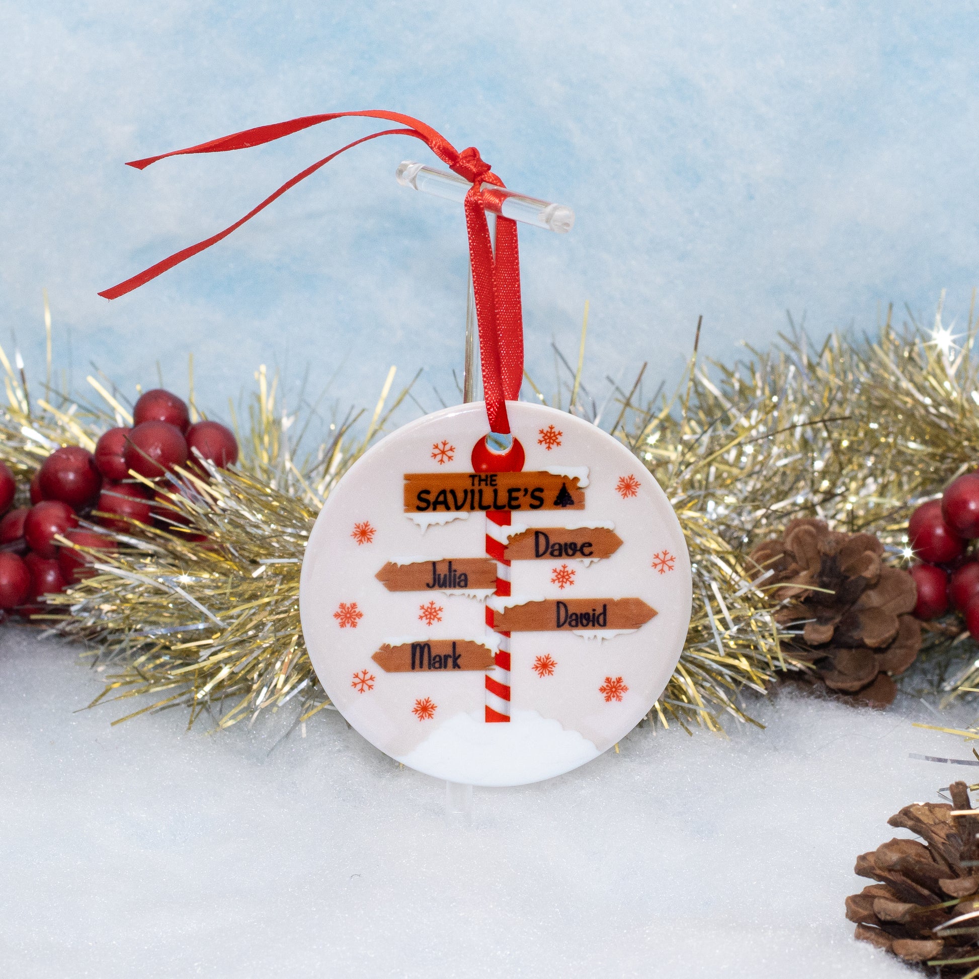 A personalized round white Christmas ornament with a red ribbon hanger, featuring printed wooden sign arrows pointing to names and text, displayed on a snowy surface with gold tinsel and red berries in the background.