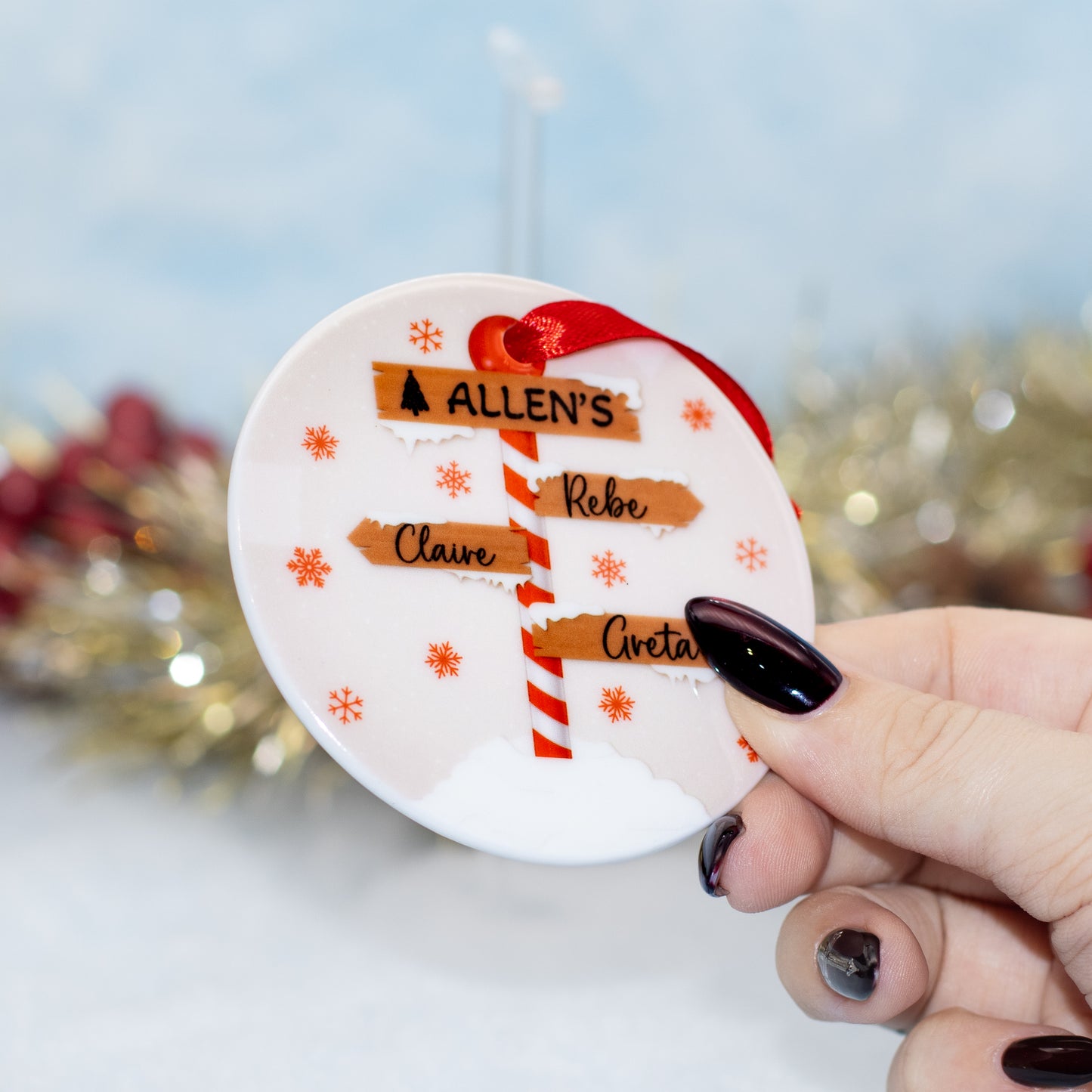 A hand holds a round white personalised Christmas ornament featuring a red-and-white striped pole with names, plus orange snowflakes and a red ribbon loop, against a softly blurred festive background.