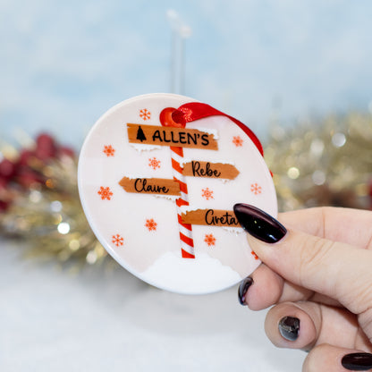 A hand holds a round white personalised Christmas ornament featuring a red-and-white striped pole with names, plus orange snowflakes and a red ribbon loop, against a softly blurred festive background.