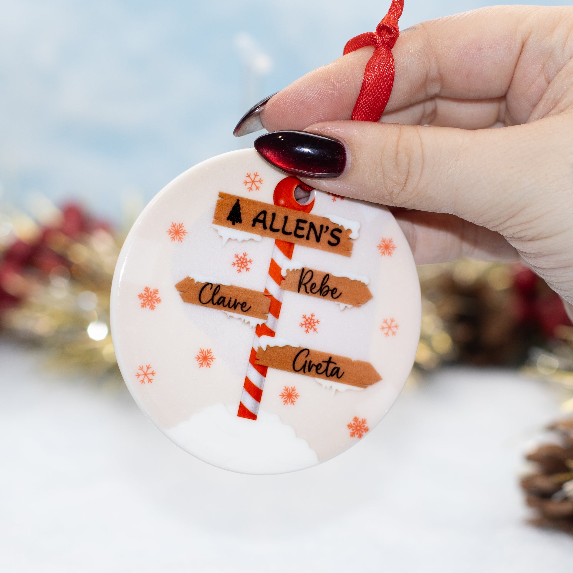 A hand holds a round white ceramic Christmas ornament featuring a red-and-white striped pole with three brown directional signs plus small orange snowflakes and a red ribbon loop at the top.