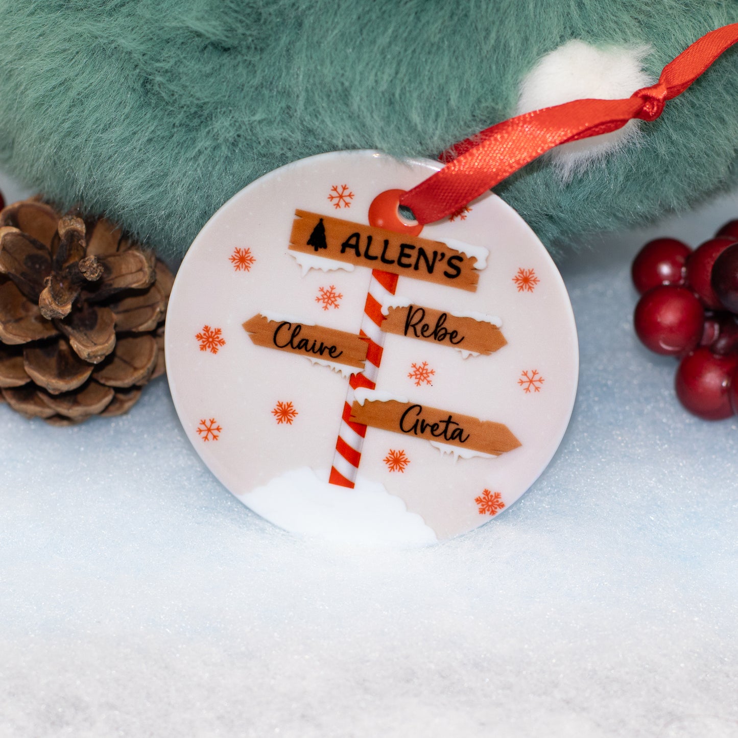 A personalised round Christmas ornament with a white background, decorated with orange snowflakes, a red-and-white striped pole,  It hangs from a red ribbon 