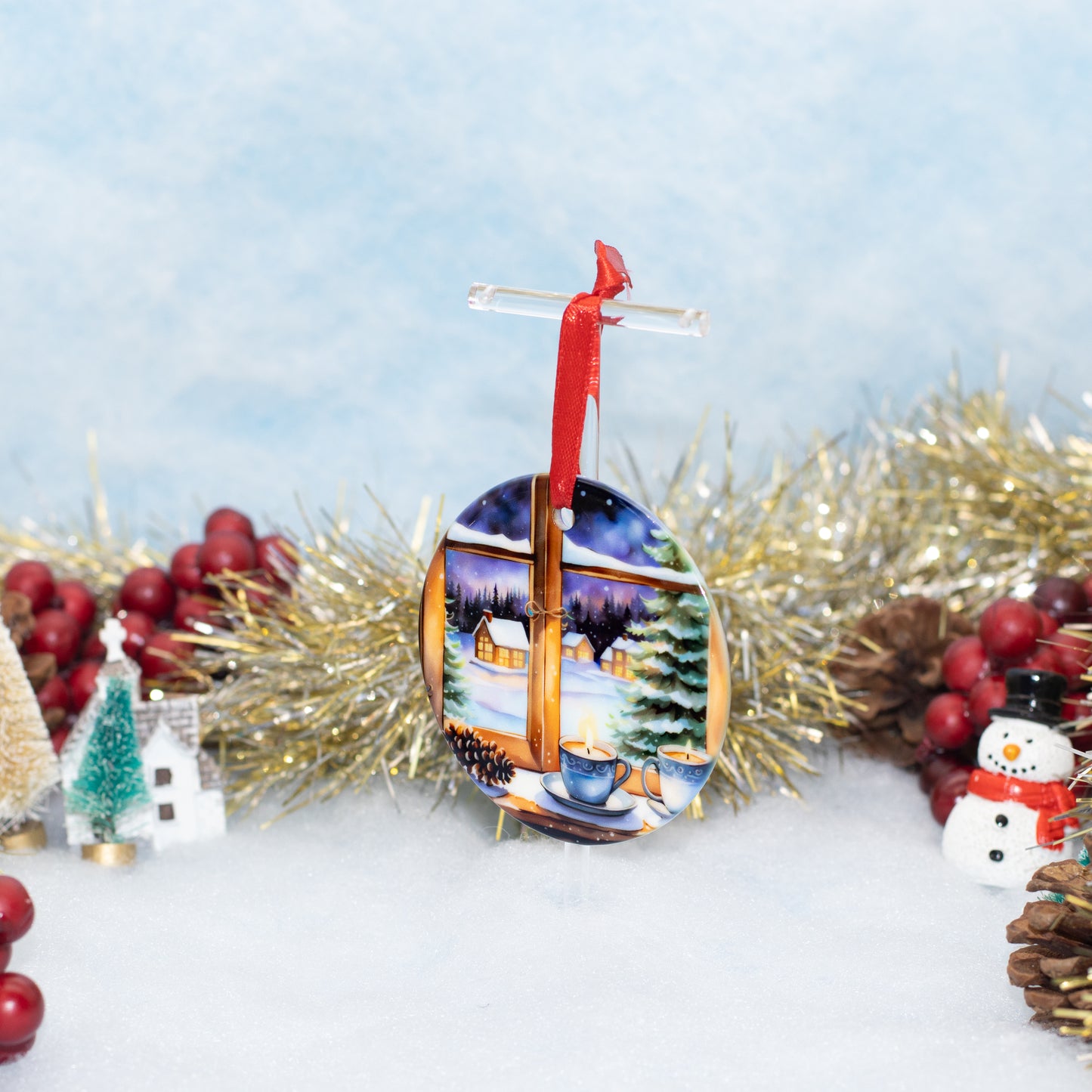 A round Christmas ornament featuring a winter village scene seen through a window, hanging from a red ribbon and clear loop, styled with gold tinsel, red berries, pinecones, and a snowman on a snowy surface against a soft blue background.