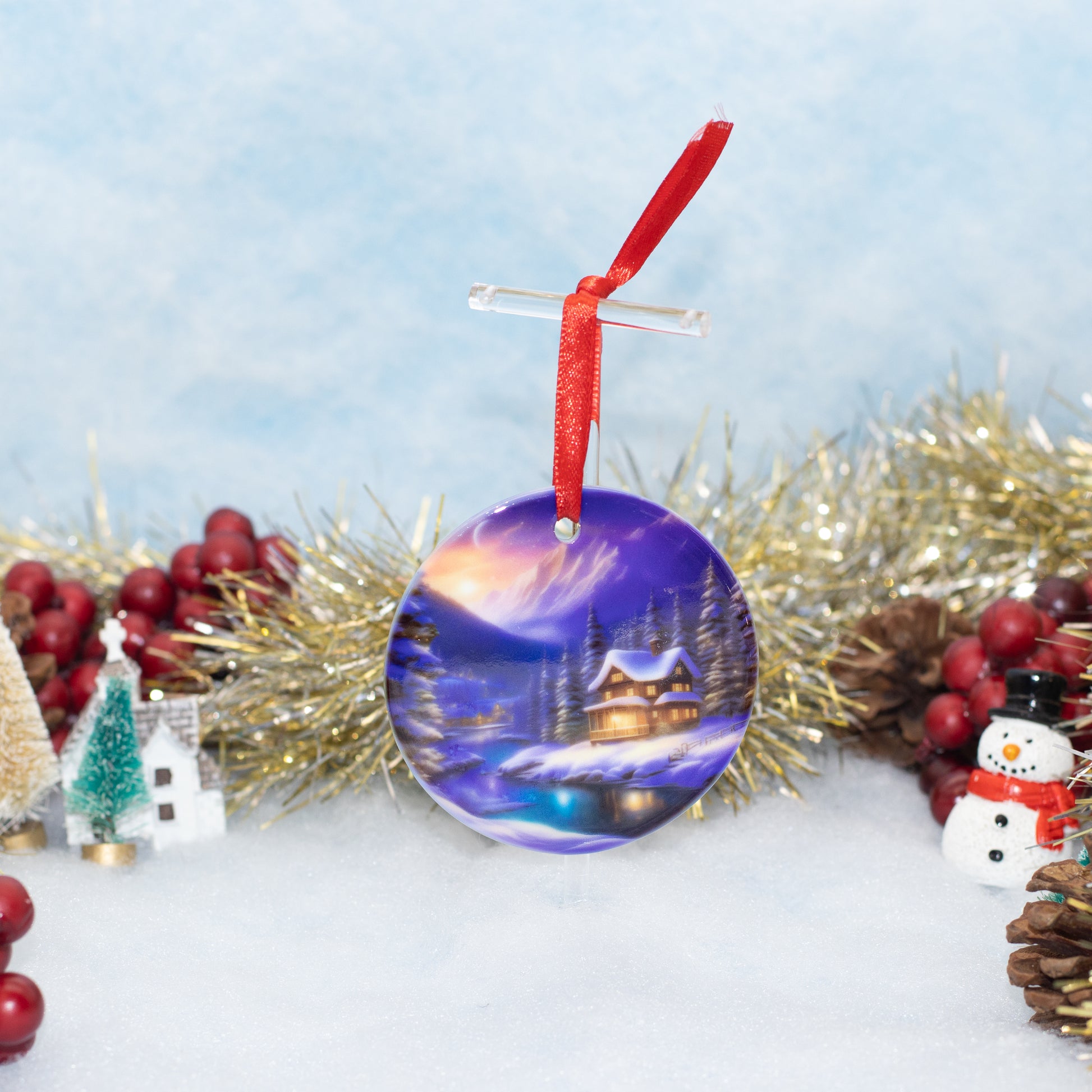 A round Christmas ornament featuring a painted winter village scene with a glowing cabin under a purple and blue night sky, hanging from a red ribbon. The ornament is styled on a snowy surface with gold tinsel, red berries, pinecones, miniature houses, and a snowman.