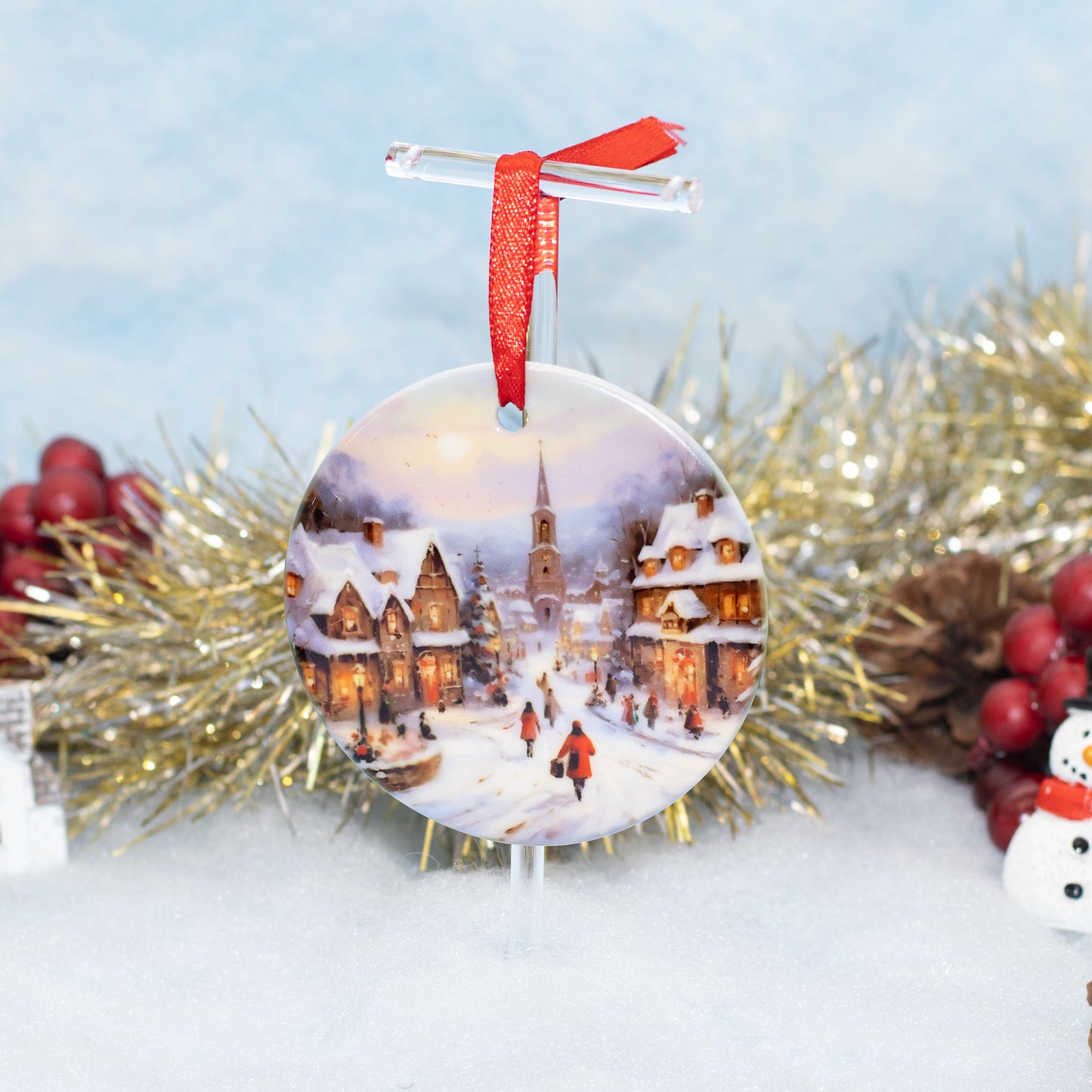 A round Christmas ornament featuring a winter village scene with snow-covered houses, warm glowing windows, and a church steeple, hanging from a clear stand with a red ribbon. The ornament is photographed in a festive setup with gold tinsel, red berries, pinecones, and a snowy surface against a soft blue background.