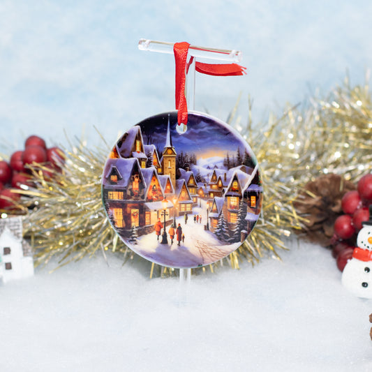 A round Christmas ornament featuring a winter village scene with glowing windows, hanging from a clear stand with a red ribbon, styled with gold tinsel, red berries, and a snowman on a snowy surface.
