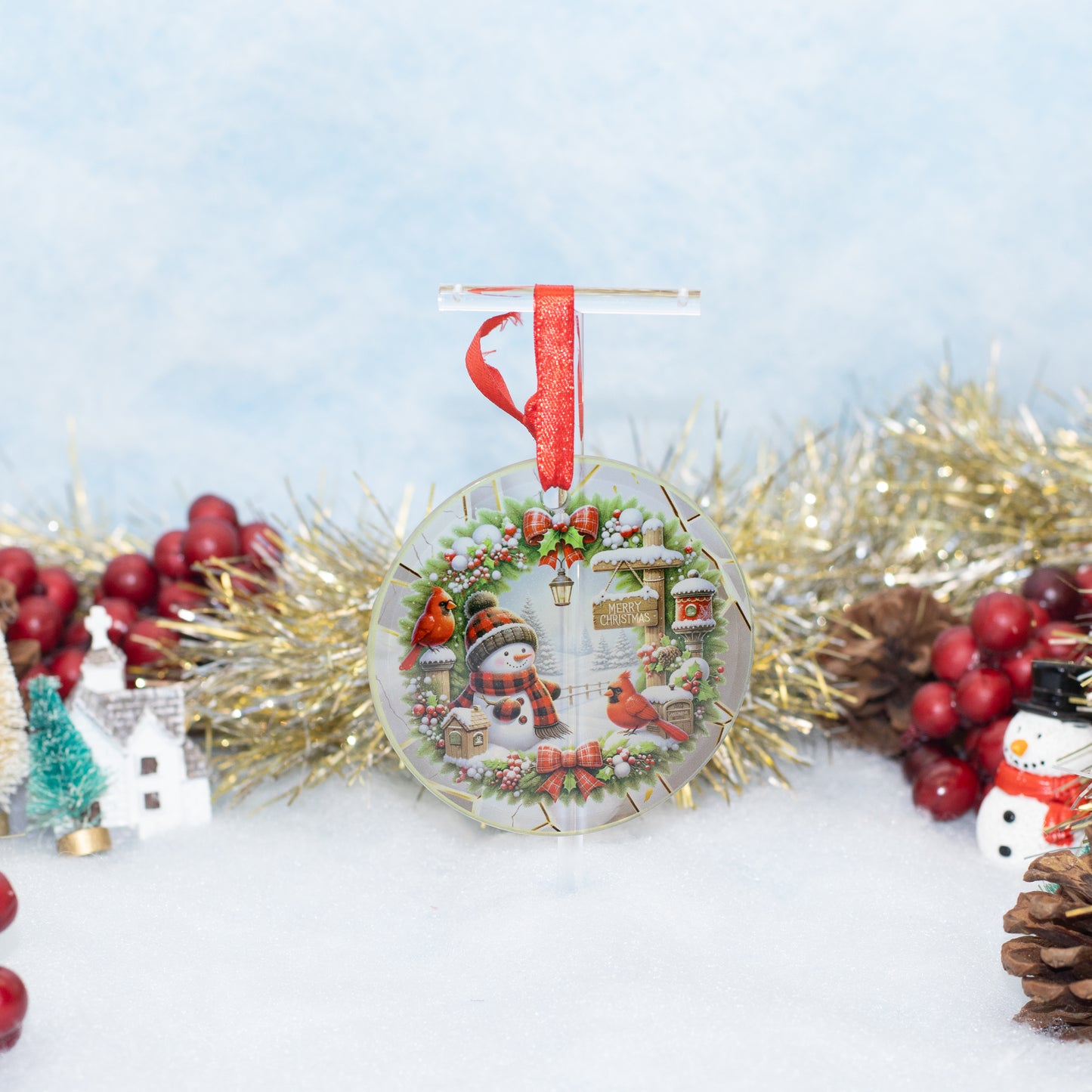 A round Christmas ornament featuring a winter village scene with a snowman, red birds, and green holly, hanging from a red ribbon. The ornament is styled on a white surface with gold tinsel, red berries, pinecones, and small holiday decorations against a soft light blue background.