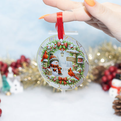 A hand holds a round clear glass Christmas ornament featuring a winter wreath scene with a snowman, birds, and red ribbon hanger against a softly blurred festive background.