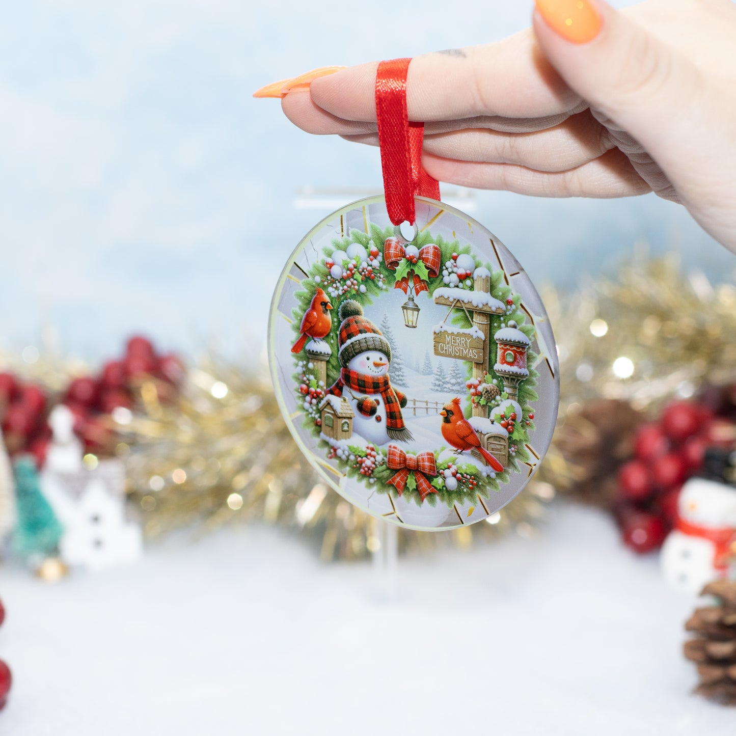 A hand holds a round Christmas ornament featuring a festive winter scene with a snowman, red cardinals, holly, and a red ribbon loop, against a softly blurred holiday background.