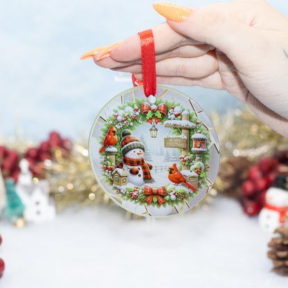 A hand holds a round glass Christmas ornament featuring a festive winter scene with a snowman, red birds, and a wreath, set against a blurred holiday background.