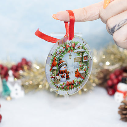 A hand holds a round clear Christmas ornament featuring a winter scene with a snowman, birds, holly, and a small house, suspended by a red ribbon against a softly blurred festive background.