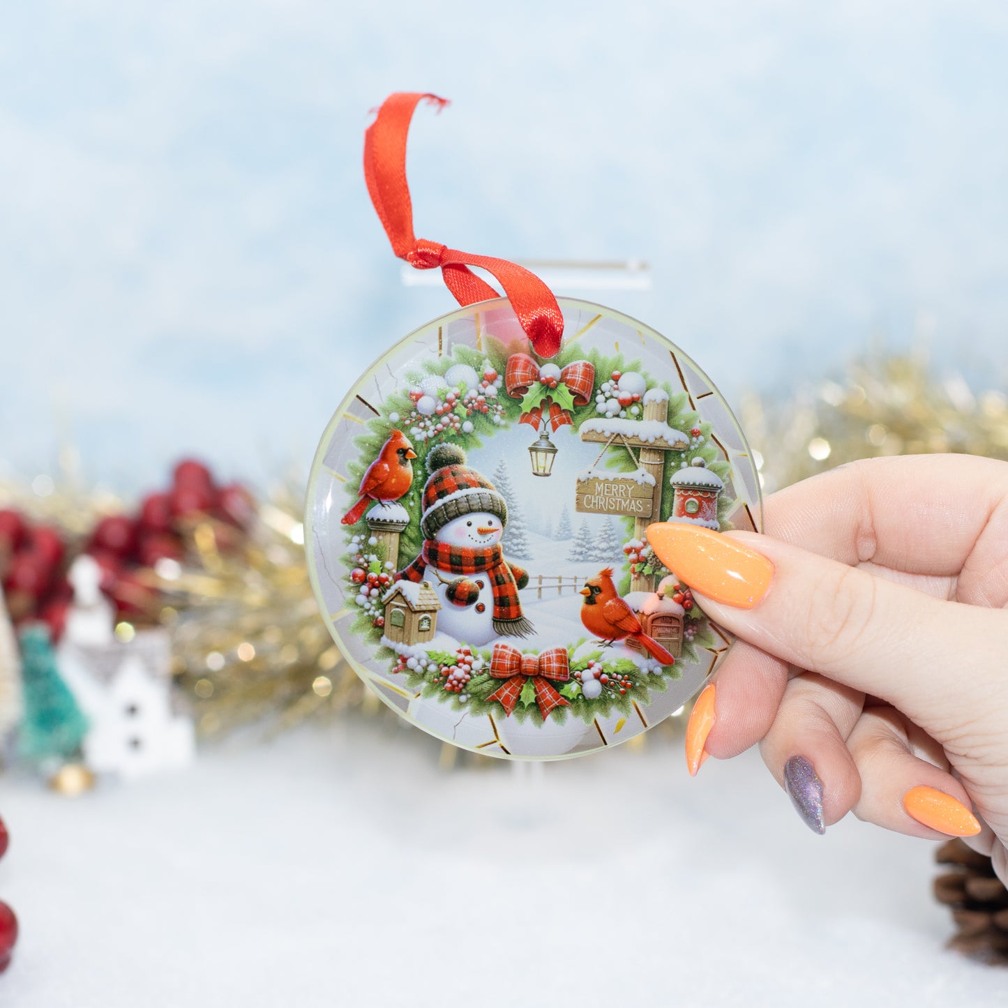 A hand holds a round clear Christmas ornament featuring a winter village scene with a snowman, red birds, and a red ribbon loop, against a softly blurred festive background.