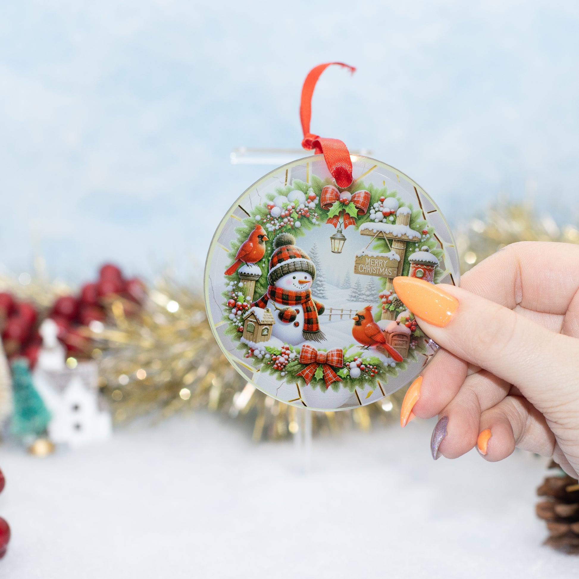 A hand holds a round clear Christmas ornament featuring a winter wreath, snowman, red birds, and small holiday cottages. The ornament has a red ribbon loop at the top and is photographed against a softly blurred festive background with gold tinsel and holiday decor.