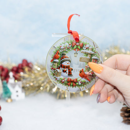 A hand holds a round clear Christmas ornament featuring a winter wreath, snowman, red birds, and small holiday cottages. The ornament has a red ribbon loop at the top and is photographed against a softly blurred festive background with gold tinsel and holiday decor.