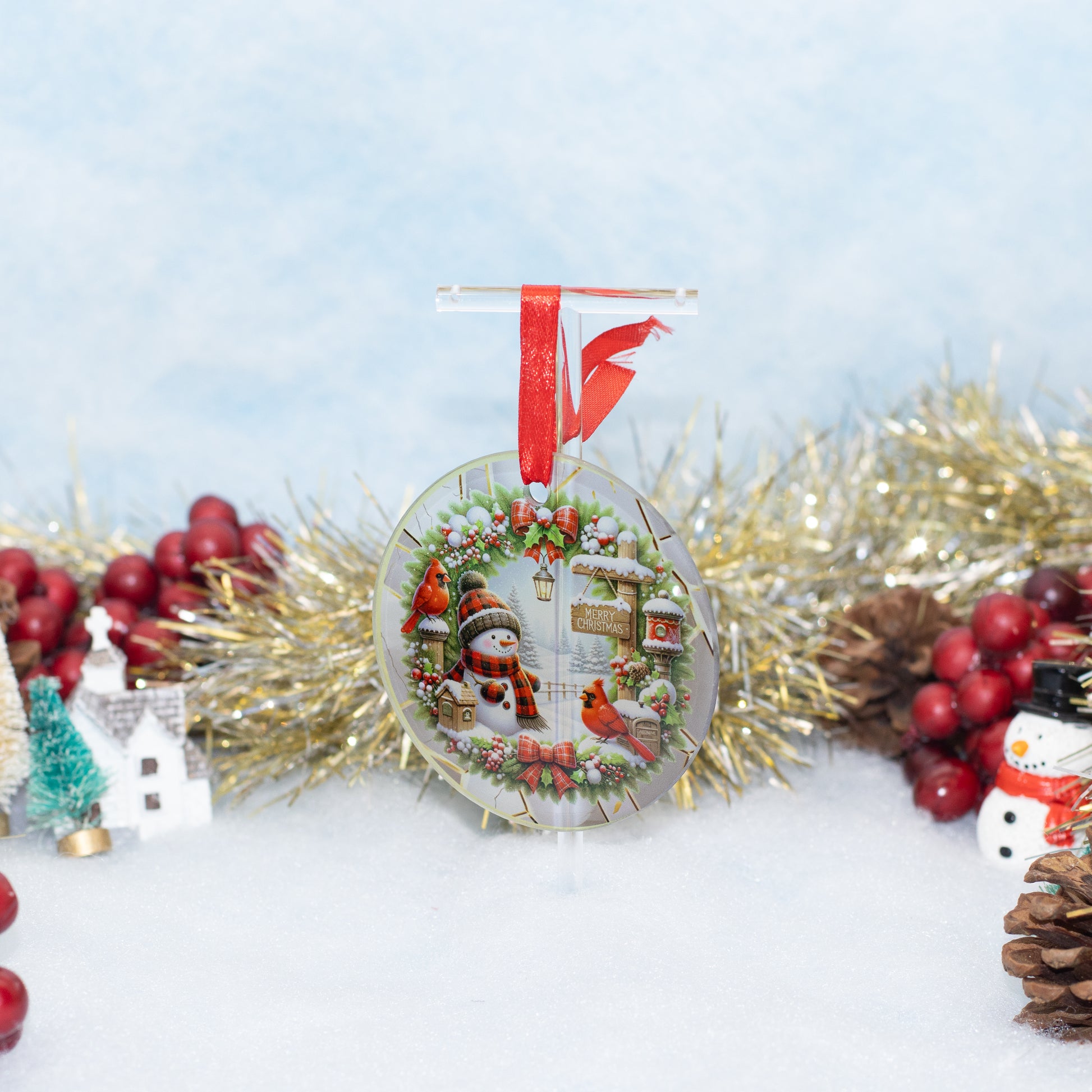 A round Christmas ornament displayed on a clear stand, featuring a festive winter scene with snowman, snowman figurines, red birds, and a wreath, hanging from a red ribbon. The ornament is styled on a white surface with gold tinsel, red berries, pinecones, and small decorative houses in the background.