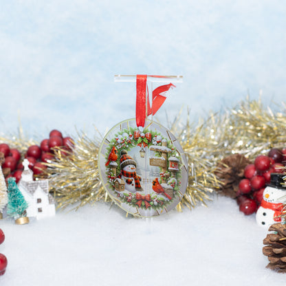 A round Christmas ornament displayed on a clear stand, featuring a festive winter scene with snowman, snowman figurines, red birds, and a wreath, hanging from a red ribbon. The ornament is styled on a white surface with gold tinsel, red berries, pinecones, and small decorative houses in the background.