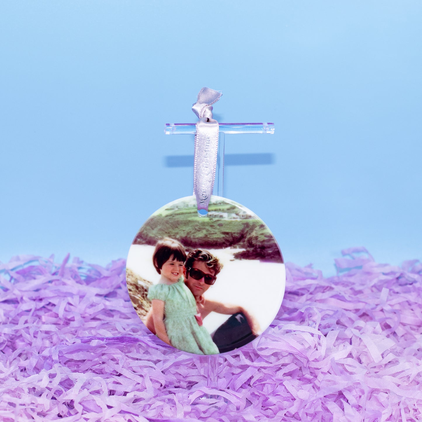 A round custom photo ornament hanging from a clear acrylic hanger with a silver ribbon, displayed on a bed of purple shredded paper against a light blue background.