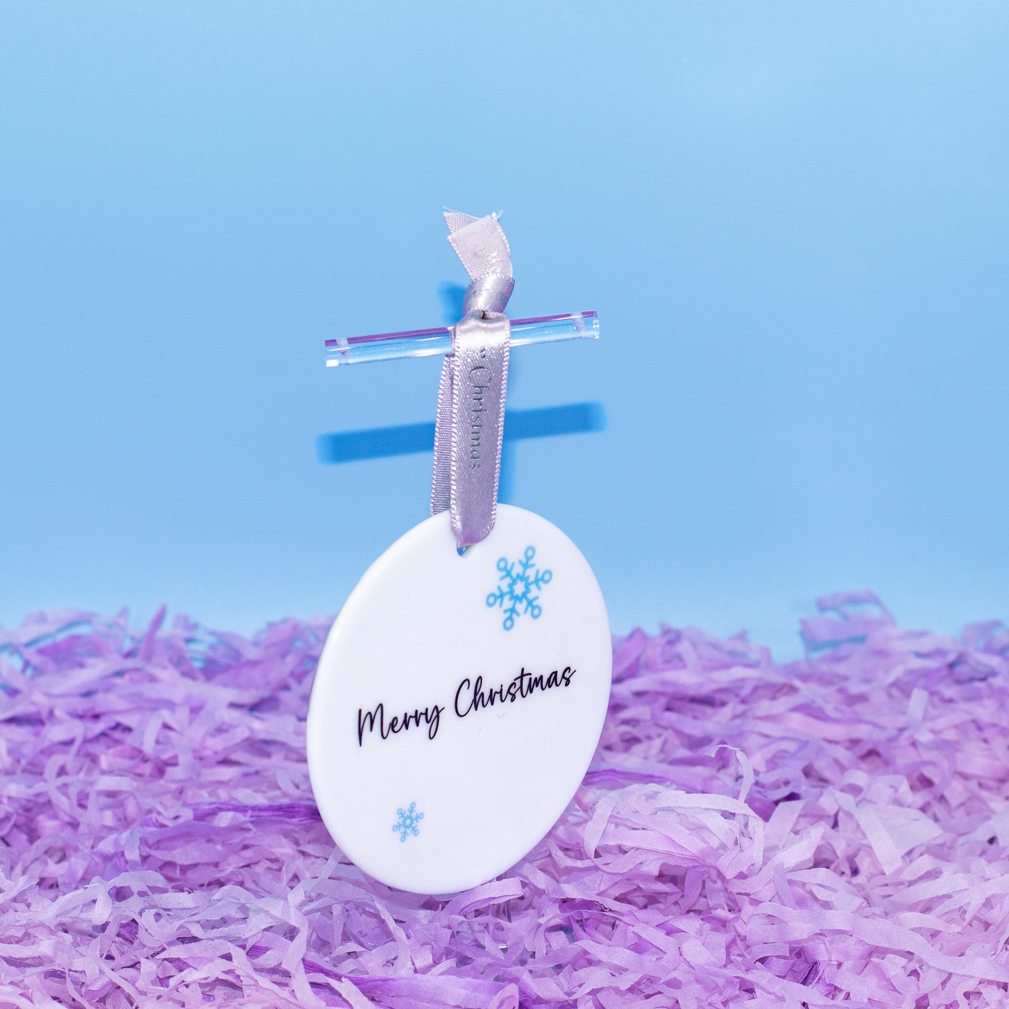 A white Christmas ornament with 'Merry Christmas' printed in black script, decorated with blue snowflakes, hanging from a silver ribbon and clear loop, standing upright in purple shredded paper against a light blue background.