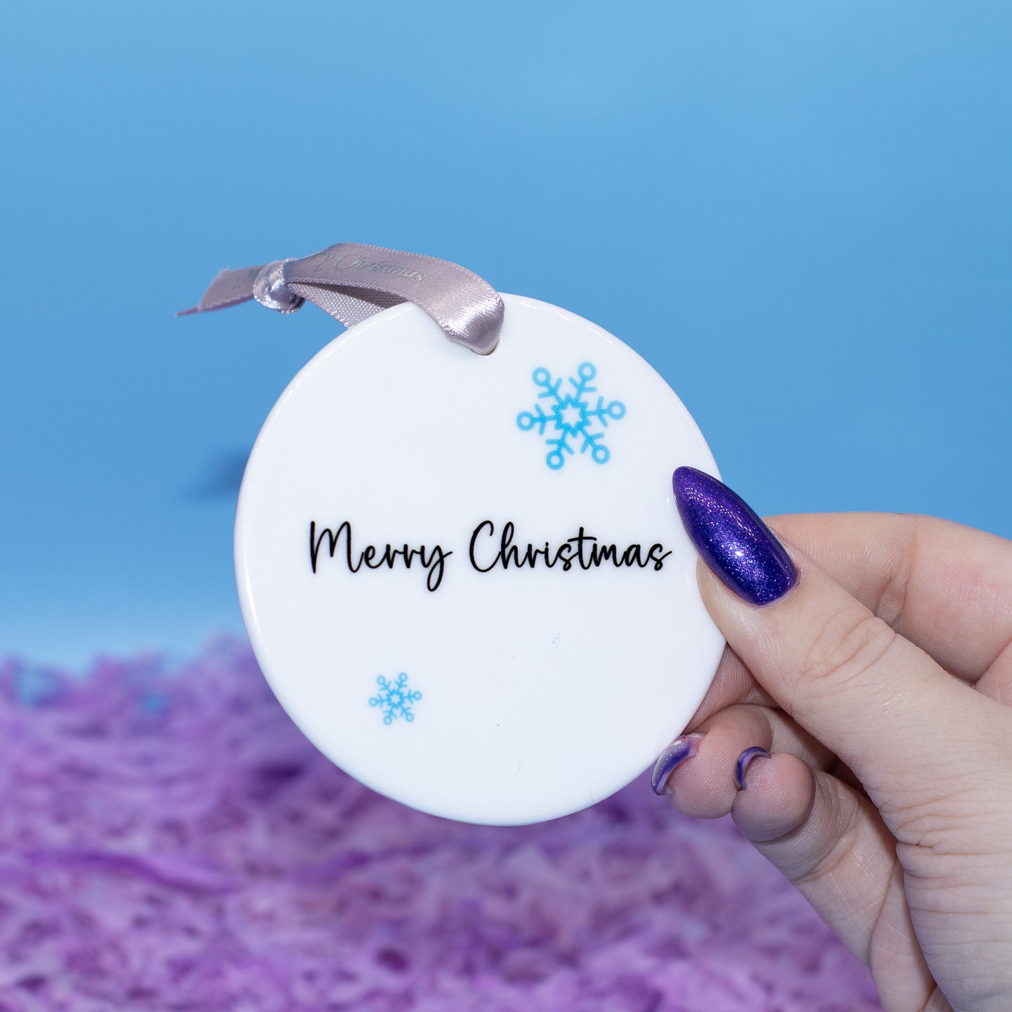 A hand holds a round white Christmas ornament that reads 'Merry Christmas' in black script, decorated with two blue snowflakes and a silver ribbon. The background is a bright blue sky with blurred purple shredded paper.