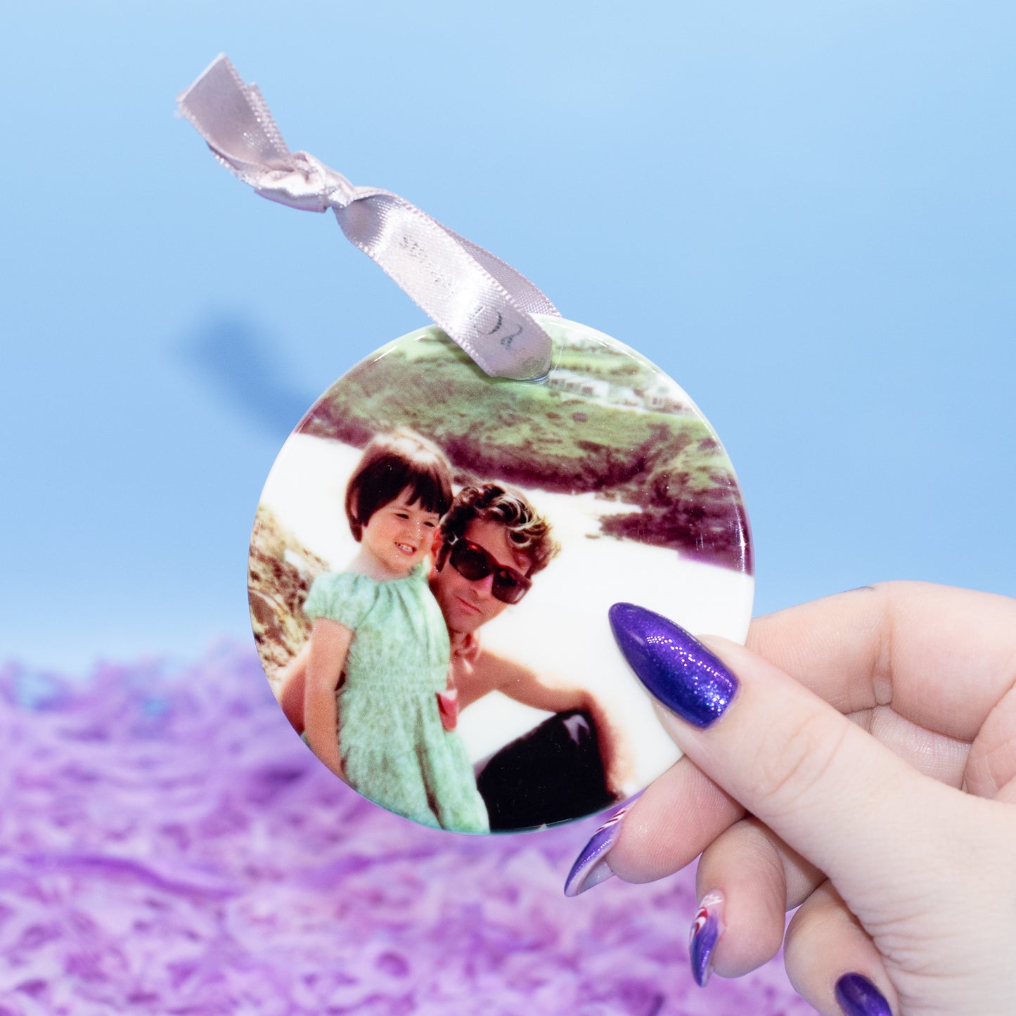 A hand holds a round custom photo ornament featuring a young girl in a green dress. The ornament has a silver ribbon loop at the top and is set against a soft-focus background of light blue and purple.