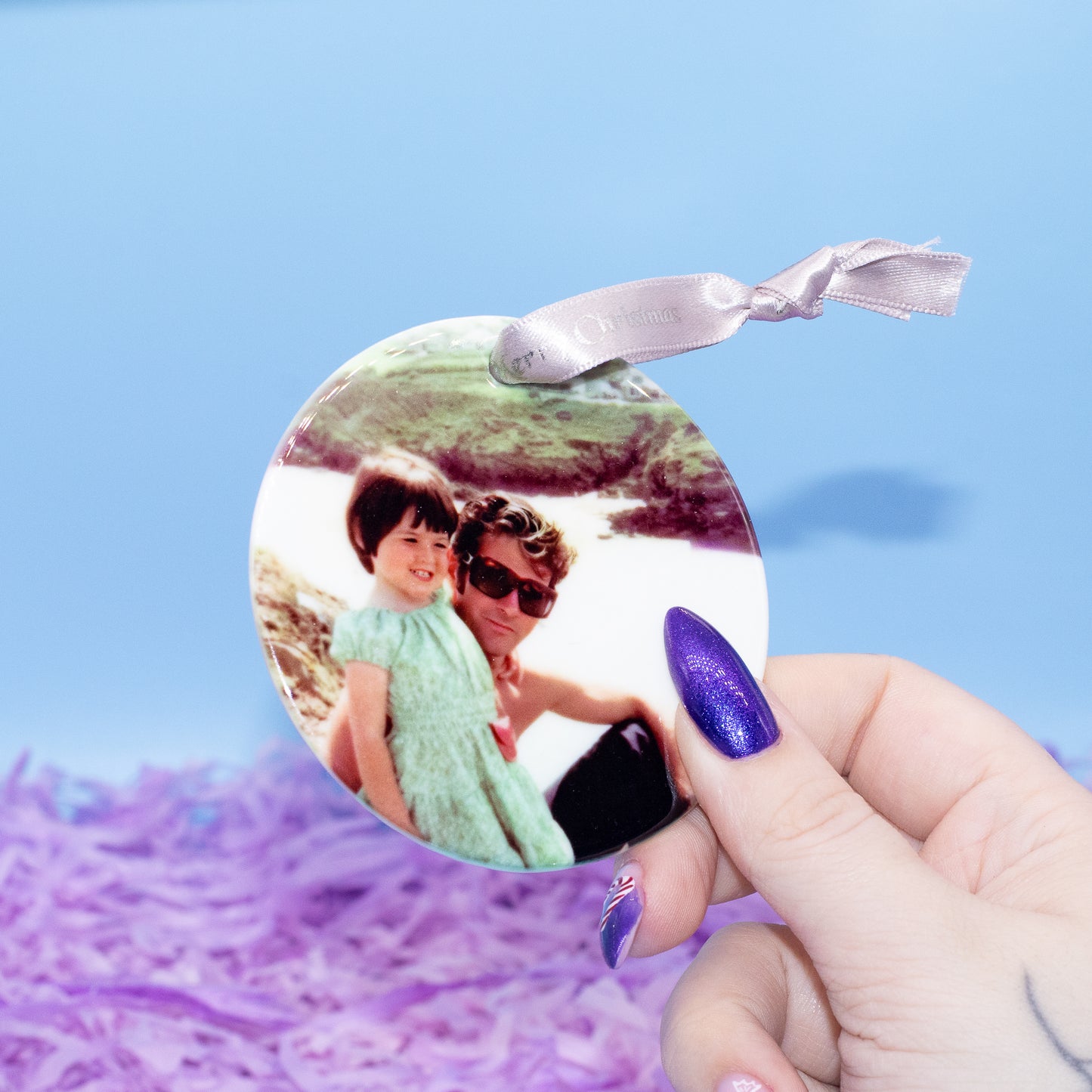 A round custom photo ornament featuring a printed photograph of a woman and child, held by a hand with purple glitter nail polish against a light blue background with purple shredded paper.