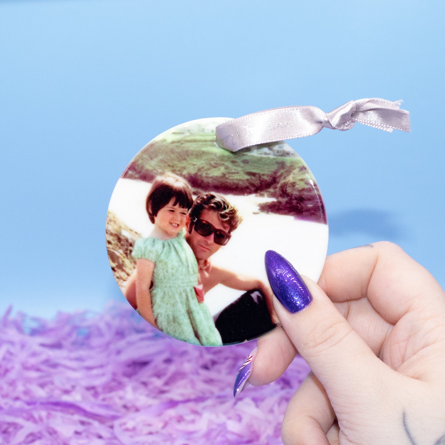 A hand holds a round custom photo ornament featuring a printed photograph of a man and child, with a silver ribbon loop at the top, against a blue background with purple shredded paper.