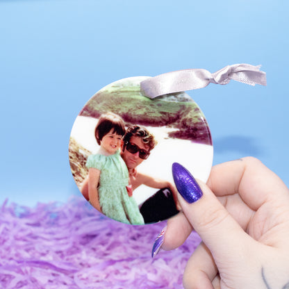 A hand holds a round custom photo ornament featuring a printed photograph of a man and child, with a silver ribbon loop at the top, against a blue background with purple shredded paper.