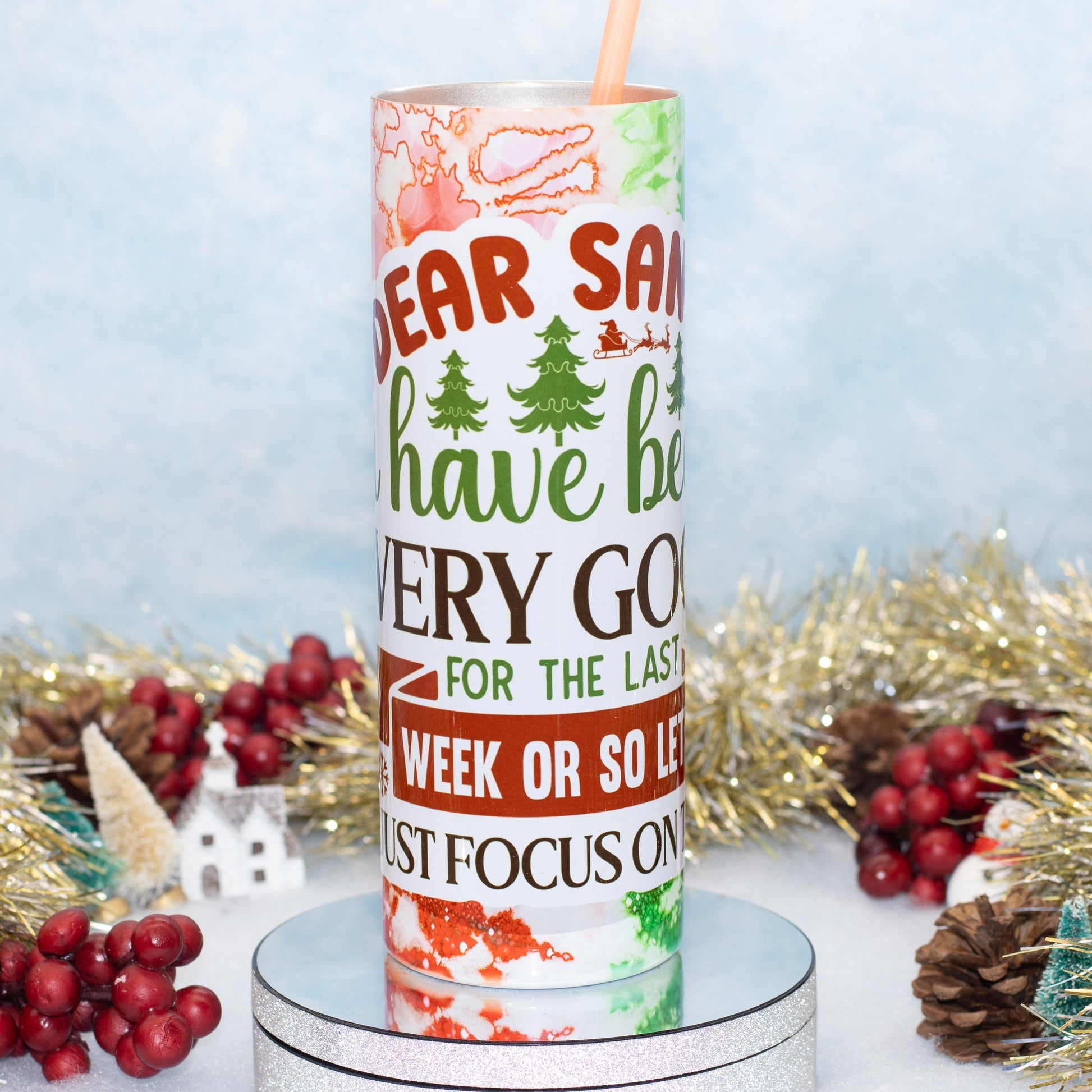 A cylindrical tumbler with a holiday-themed print reading 'Dear Santa, have been very good for the last week or so let's just focus on' in red, green, and brown lettering, shown on a silver pedestal with festive tinsel, pinecones, and red berries around it.