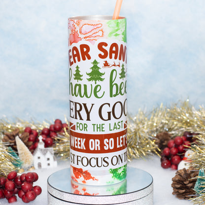 A tall insulated tumbler with a straw, decorated with a Christmas-themed graphic that reads 'Dear Santa have been very good for the last week or so let's just focus on this week,' displayed on a silver pedestal with holiday decorations in the background.