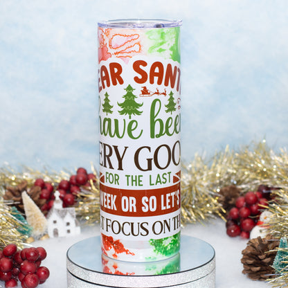 A tall insulated tumbler with a festive Christmas design featuring the text 'Dear Santa have been very good for the last week or so let's just focus on the next week' in red, green, and brown lettering. The tumbler is displayed on a silver round pedestal with holiday decorations like gold tinsel, pinecones, and red berries in the background against a light blue textured backdrop.