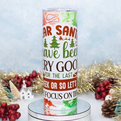 A cylindrical tumbler with a festive Christmas design featuring the text 'Santa have been very good for the last week or so let's focus on the' in red, green, and black lettering, displayed on a silver pedestal with holiday decorations in the background.