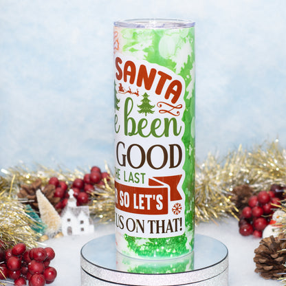 A tall insulated tumbler with a festive Christmas design, The tumbler is displayed on a silver pedestal with holiday decorations like red berries, gold tinsel, and a small white house in the background.