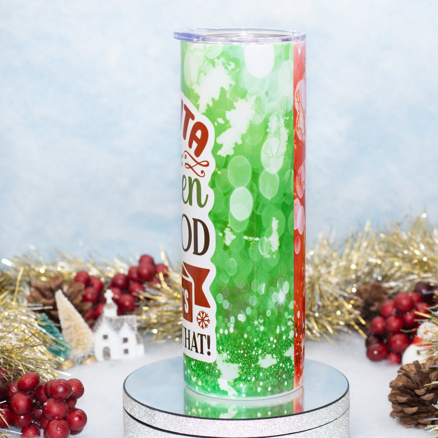 A tall tumbler with a printed green holiday design featuring white snowflakes, red text, and a red side panel, displayed on a round silver pedestal with Christmas decorations in the background.