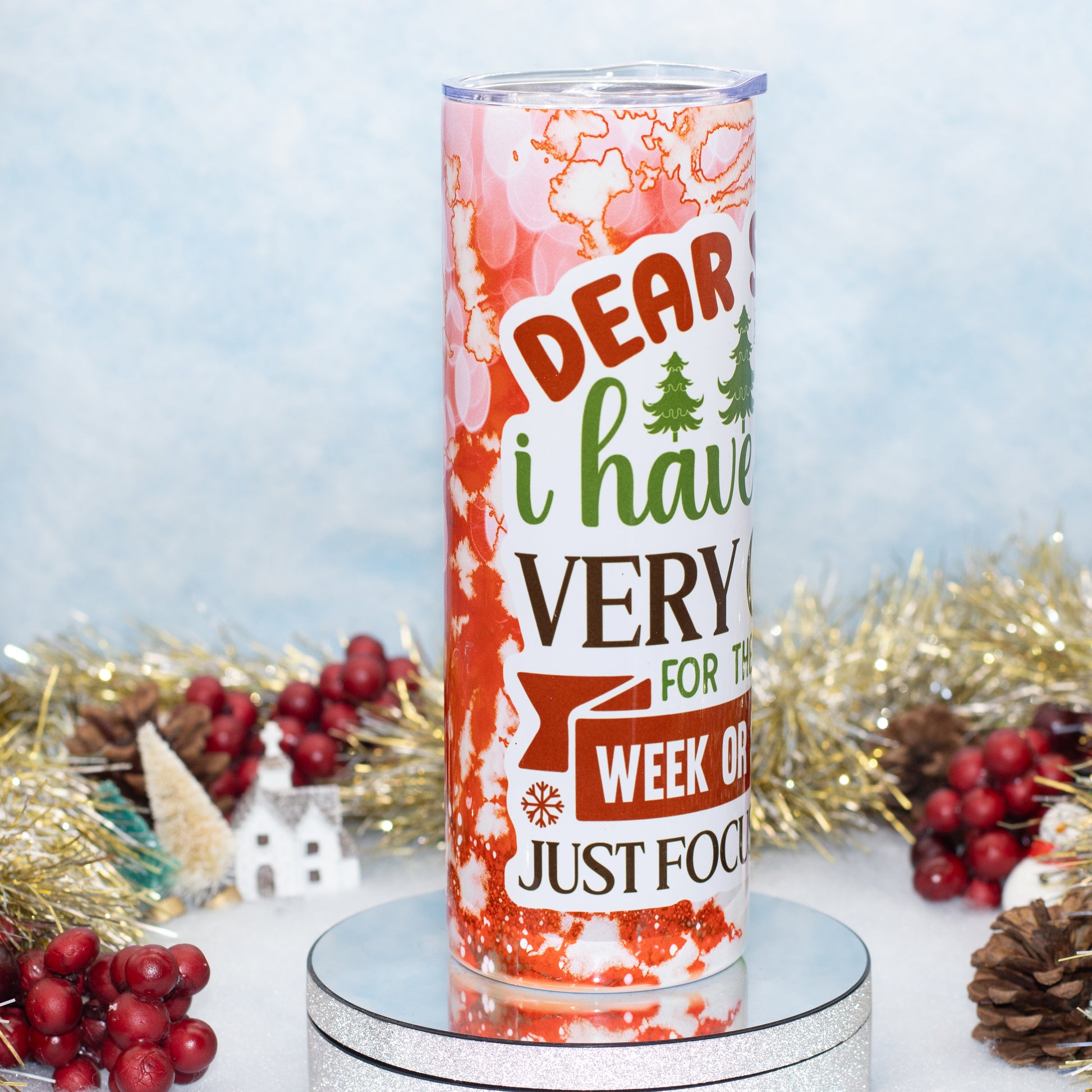 A stainless steel tumbler with a printed holiday message surrounded by festive tinsel, red berries, and pinecones on a light blue background.