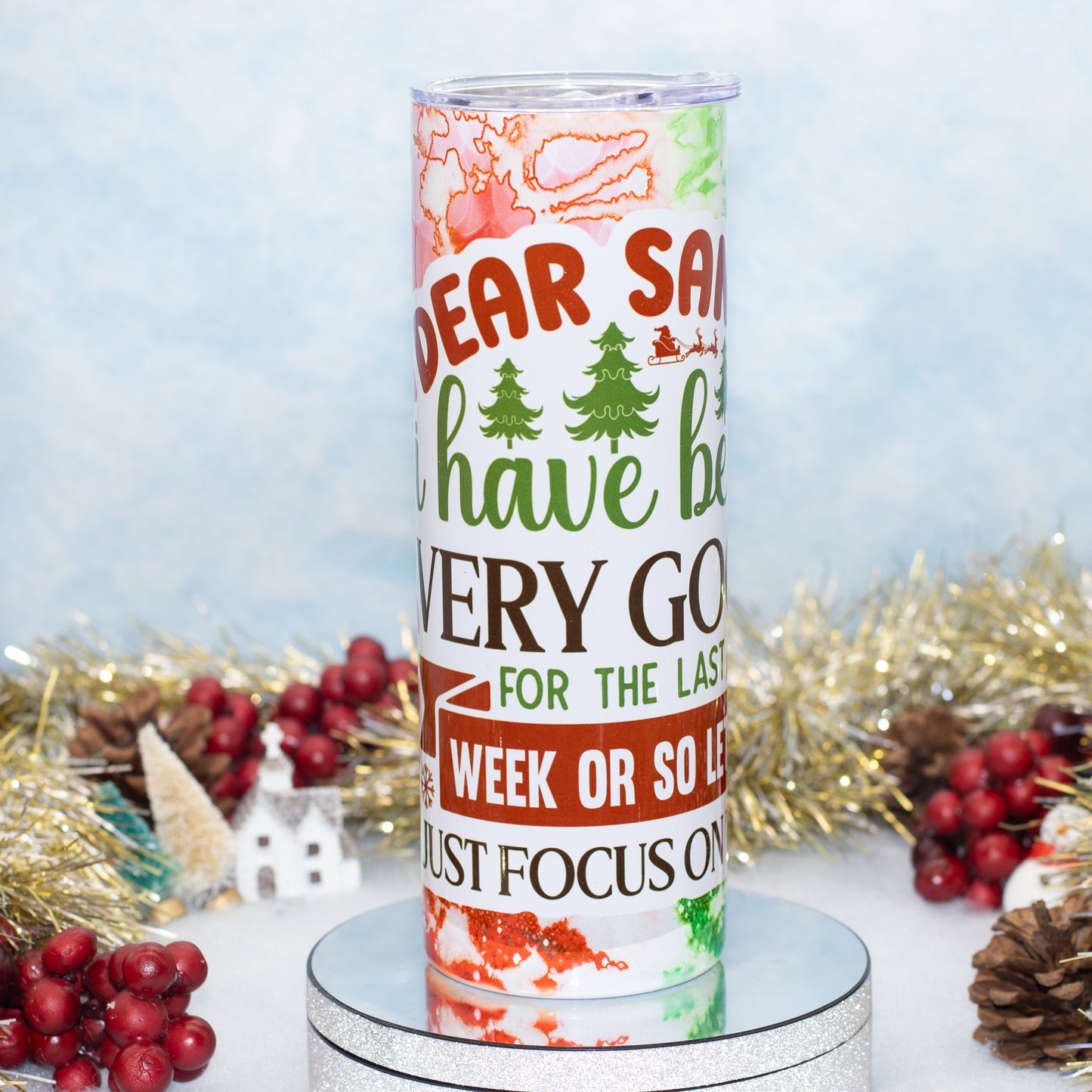 A tall insulated tumbler with a festive Christmas design, featuring a printed message about 'Dear Santa' on a white background with red, green, and brown holiday graphics. The tumbler is displayed on a round mirrored pedestal, surrounded by holiday decorations including gold tinsel, pinecones, and red berries against a light blue textured backdrop.