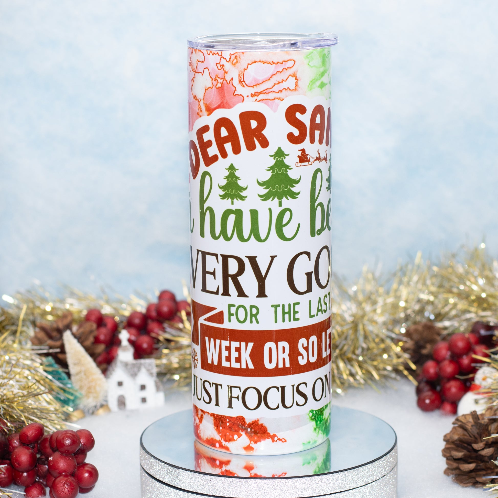 A tall insulated tumbler with a festive Christmas design, featuring a printed message about 'Dear Santa' on a white background with red, green, and brown holiday graphics. The tumbler is displayed on a round mirrored pedestal, surrounded by holiday decorations including gold tinsel, pinecones, and red berries against a light blue textured backdrop.